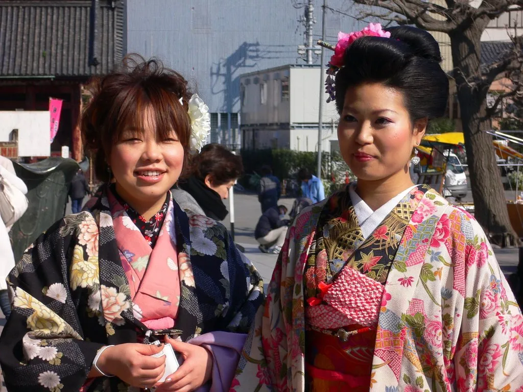 Two young women in ornate floral kimono on a sunlit street in Japan, one wearing traditional bridal hair styling with pink flowers.