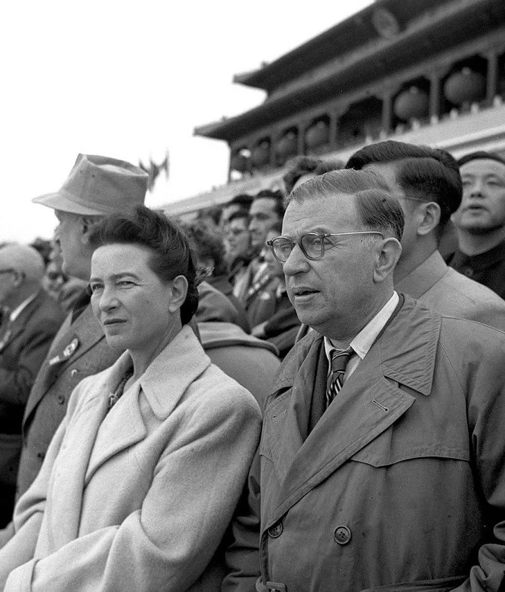 Black-and-white photograph of Simone de Beauvoir and Jean-Paul Sartre seated side by side in a crowd at a public event in Beijing, 1955.
